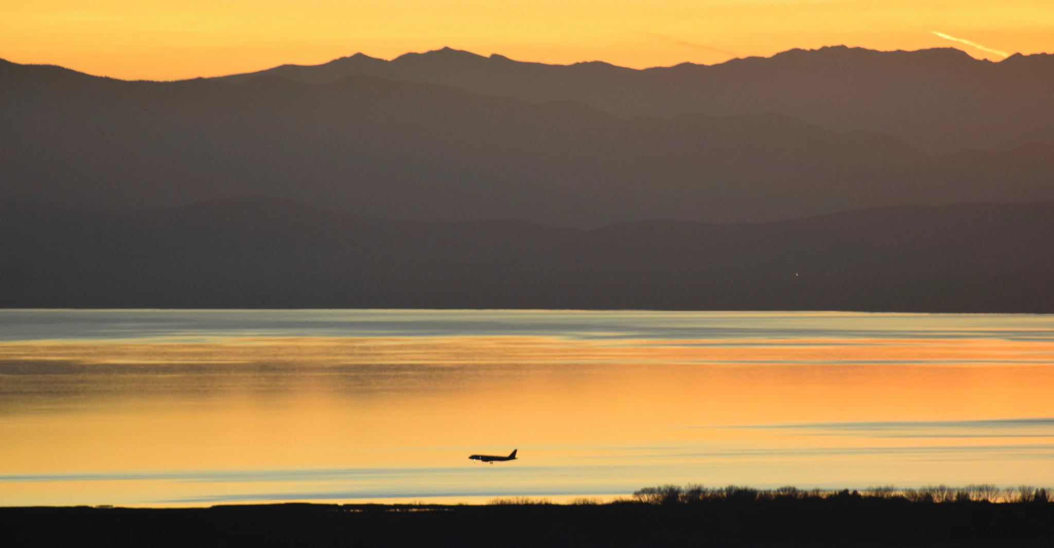 A plane flies low over a lake, which reflecs the orange sky of a sunset. The lake is backed by tall mountains which are given depth by the end of the day’s haze.