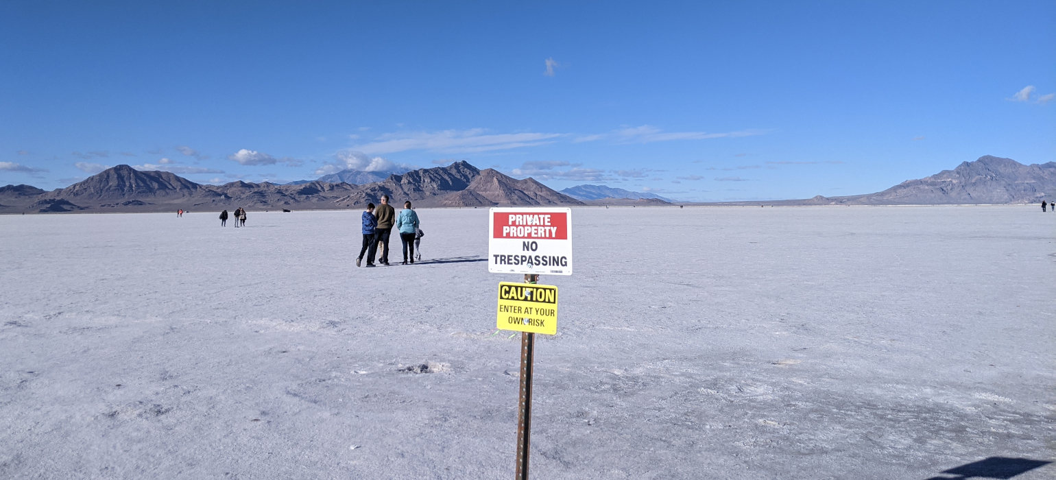 Salt flats with no trespassing sign in foreground, people beyond it, and mountains in the background