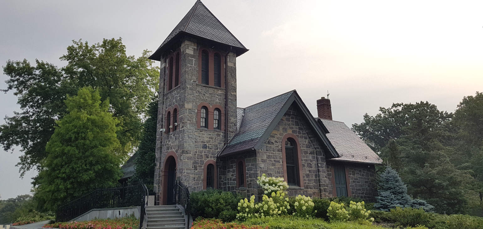 Stone building with arched windows, a tower, steps leading up, and lush lawn, flowers, and trees