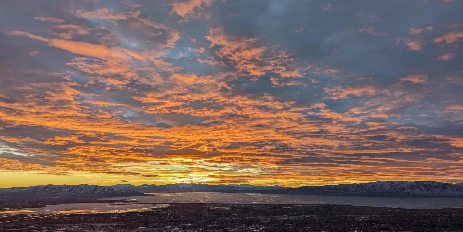 Sunset view from mountain height overlooking valley with city, lake, mountains with some snow, below yellow horizon and orange clouds under a blue sky