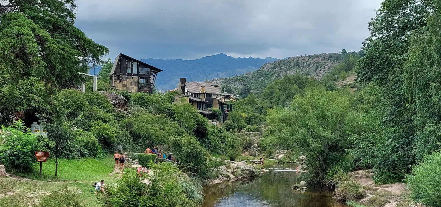 Beautiful cloudy mountain scene with river flowing by lush banks with people swimming, relaxing, and walking towards multistory buildings