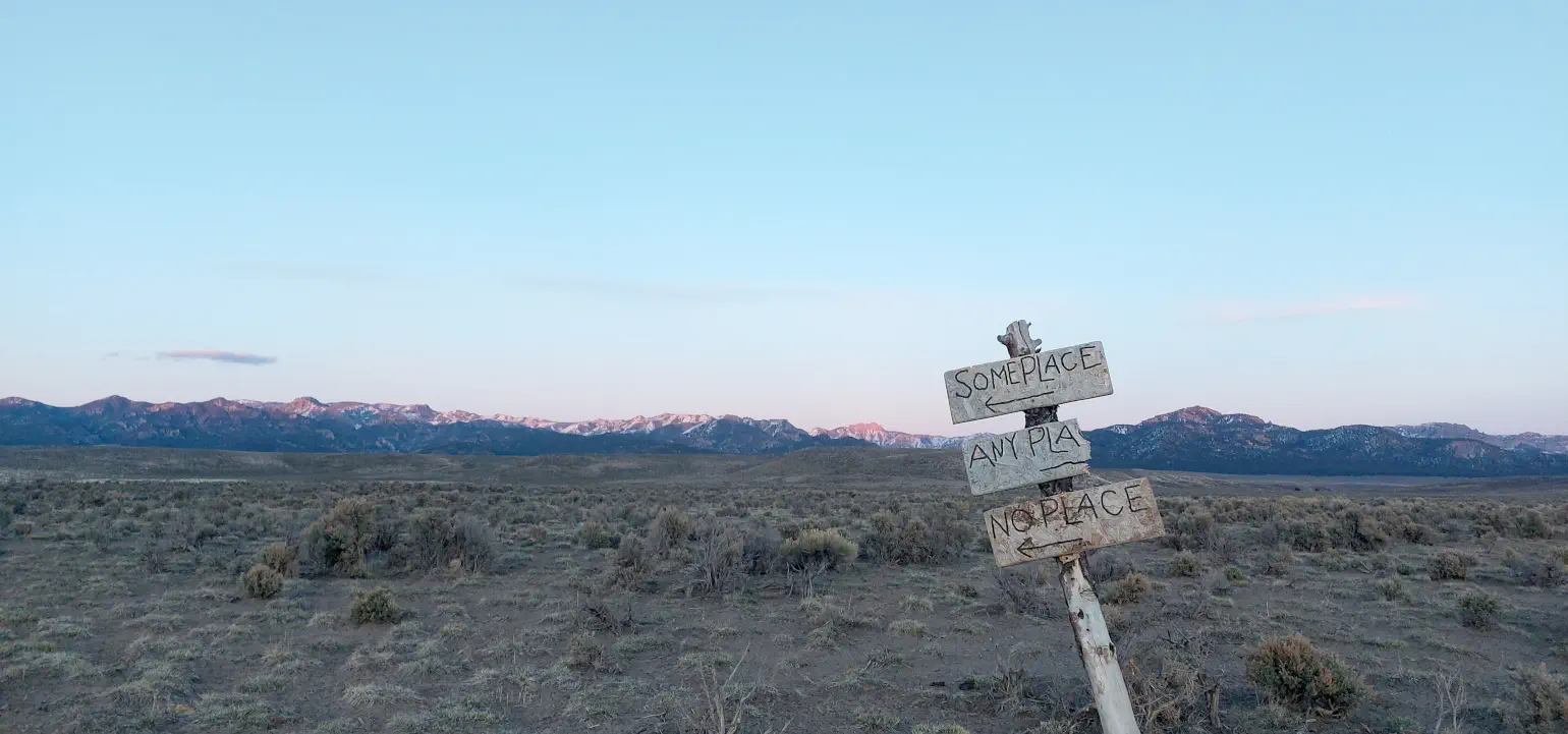 Hand-drawn signs reading “Someplace”, “Any pla…”, “No place”, with arrows pointing variously, attached to a leaning signpost in front of a high mountain desert scene with snow-topped peaks and sagebrush
