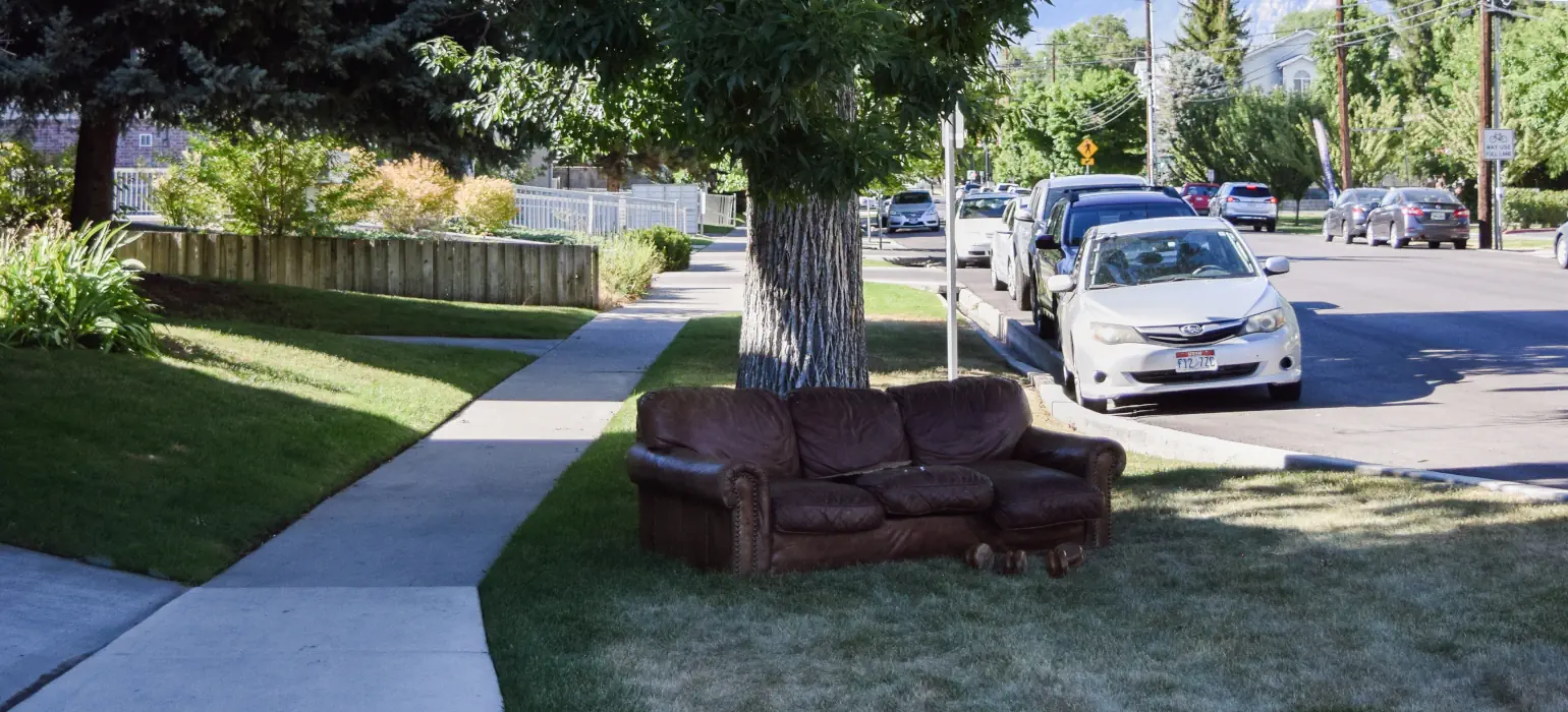 A leather couch in surprisingly good condition sits on a patch of grass between the sidewalk and the road. Harsh sunlight casts shadows of trees and buildings on the street and couch.