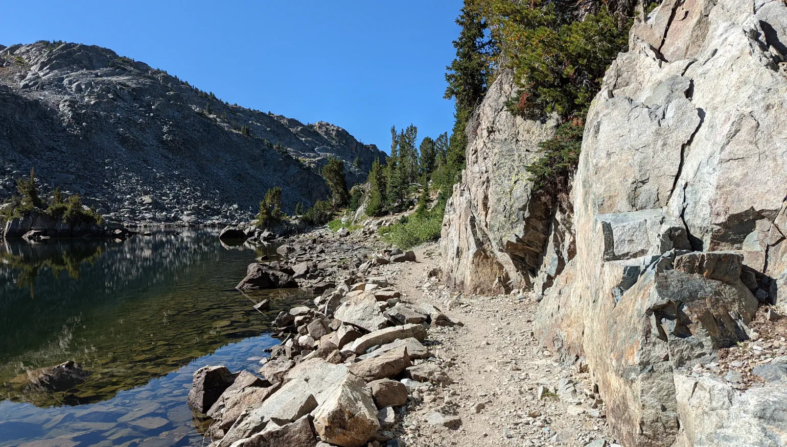 Photo of a wilderness footpath over stone with scattered evergreen trees alongside a clear lake, with a blue sky