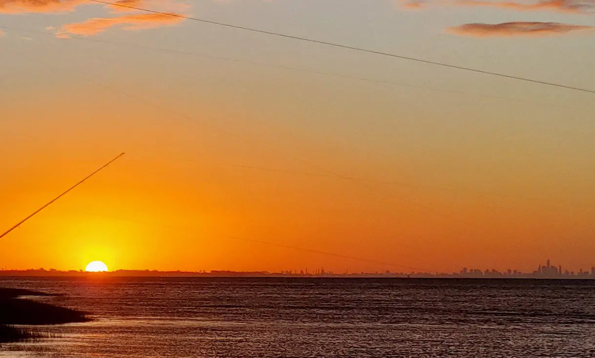 An orange sunset reflecting off of the sea at Rio de la Plata. A city skyline is silhouetted by the sunset.