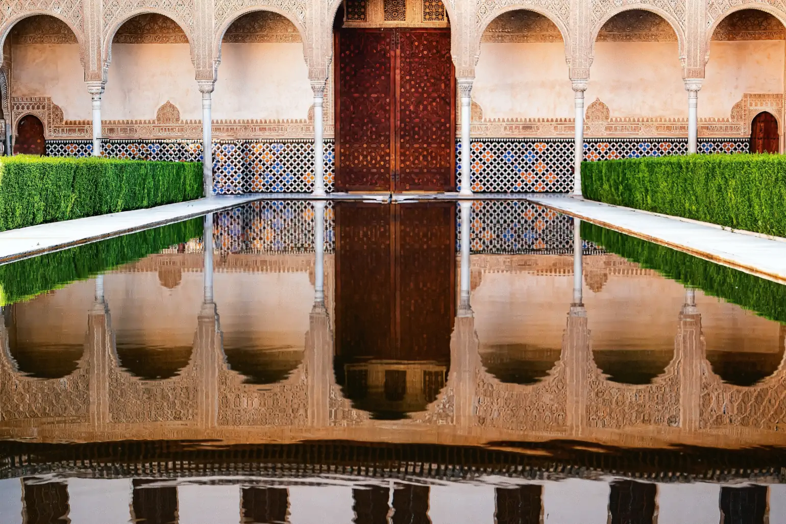 The door of Alhambra Palace, Spain. A still pool reflects grand doors, flanked on each side by arches and hedges.