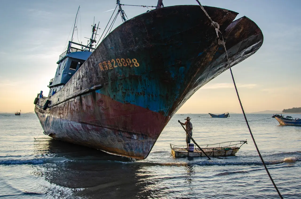 A docked fishing ship faces the camera. A man stands on a dinghy next to it.
