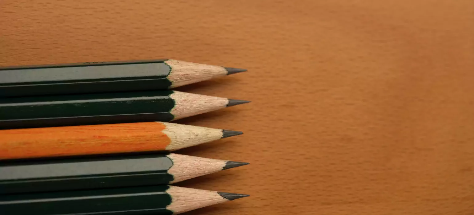 Close up photo of 5 pencils on a faux wood grain desk. The center pencil’s coating is a bright orange, while the other four are a dark green, almost black.