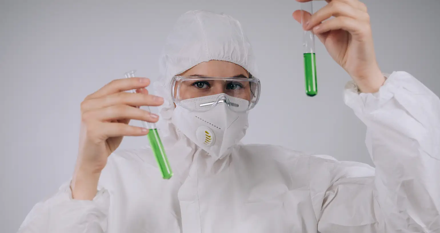 A chemist in complete PPE holds two test tubes holding green liquid.