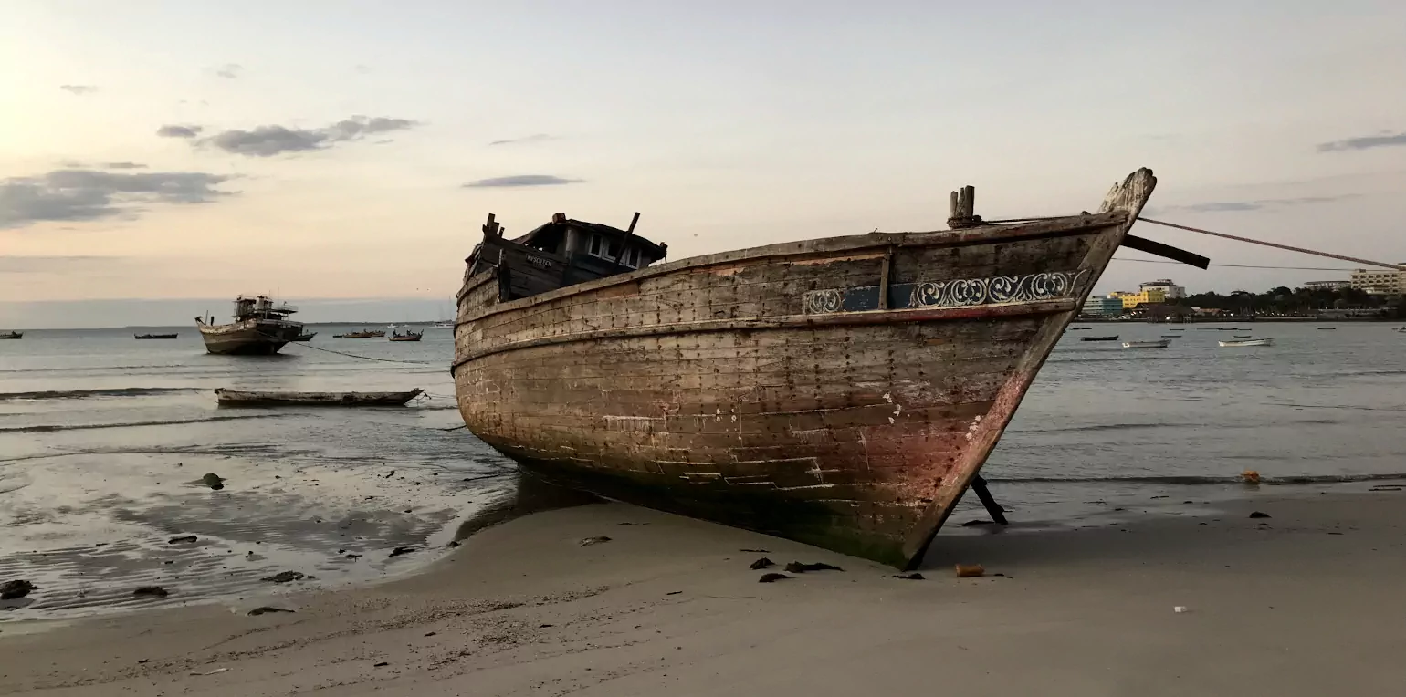 Fishing boat in Dar es Salaam. A traditional fishing boat sits on the beach at low tide, with the fading light of sunset behind. In the background, other boats float on the Msasani Bay, and several high-rise buildings are visible to the right on the Masaki peninsula.