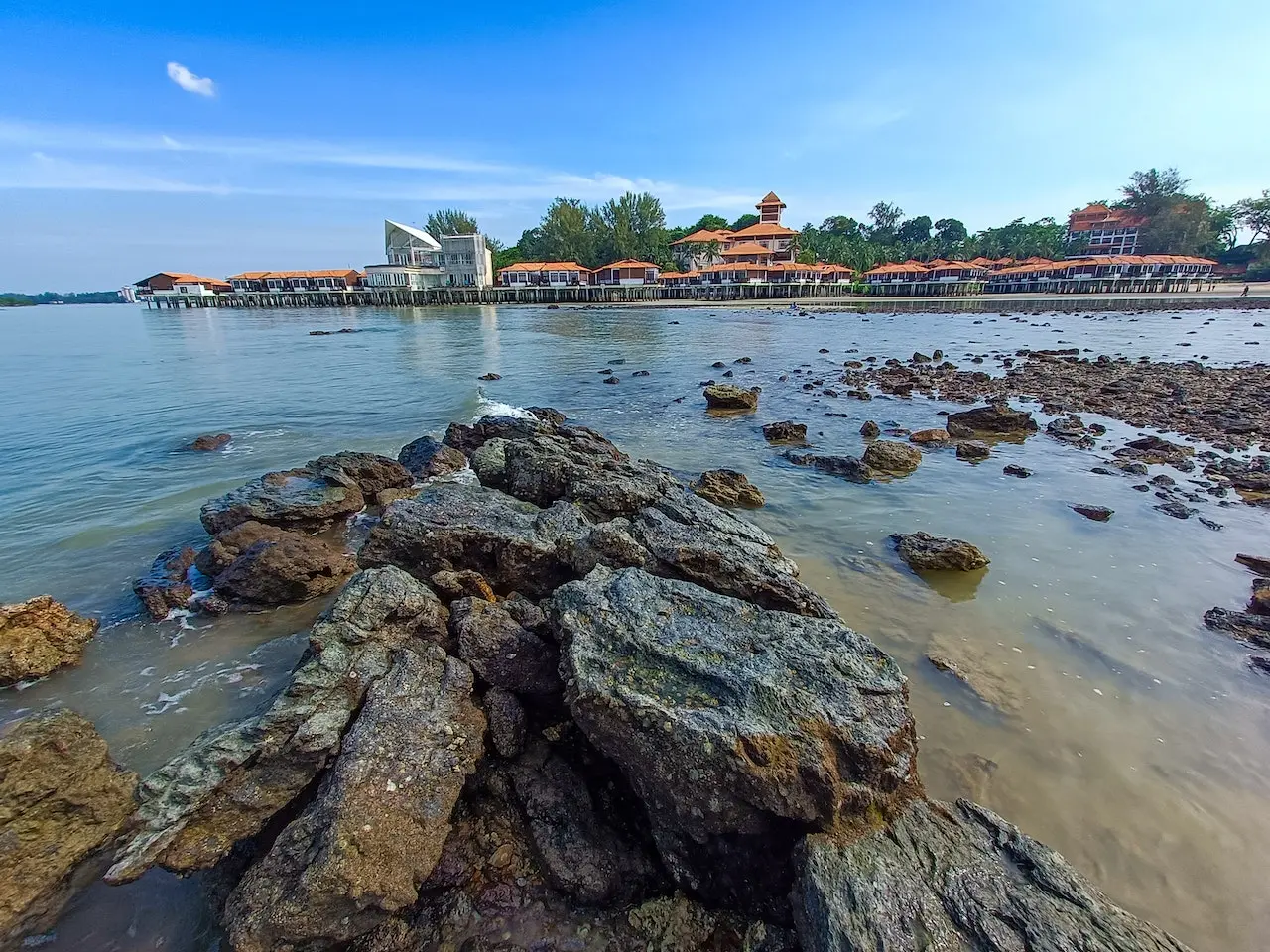 Port Dickson, a Malaysian Beach. Rocks in the forground jut out into an inlet, across which is a line of red-roofed houses.
