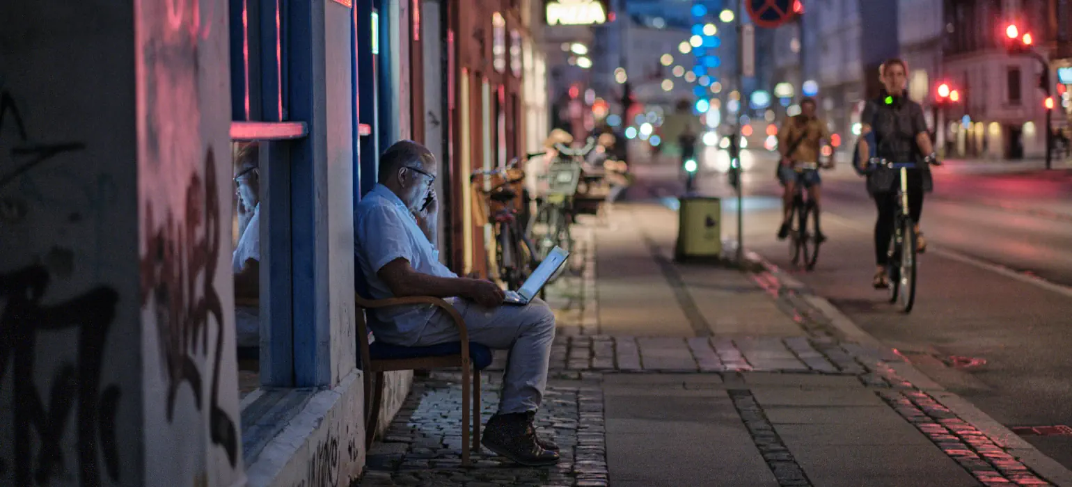 A city street at night. A man sits on a bench, looking at his laptop as cyclists pass by.