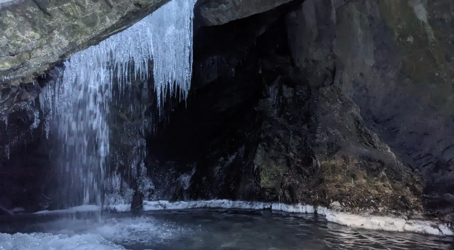 Icicles hang down from the opening of a cave, amid water falling into a pool lined with thick ice. Light from the cave’s opening illuminates the bottom corner of the image, opposite the icicles.