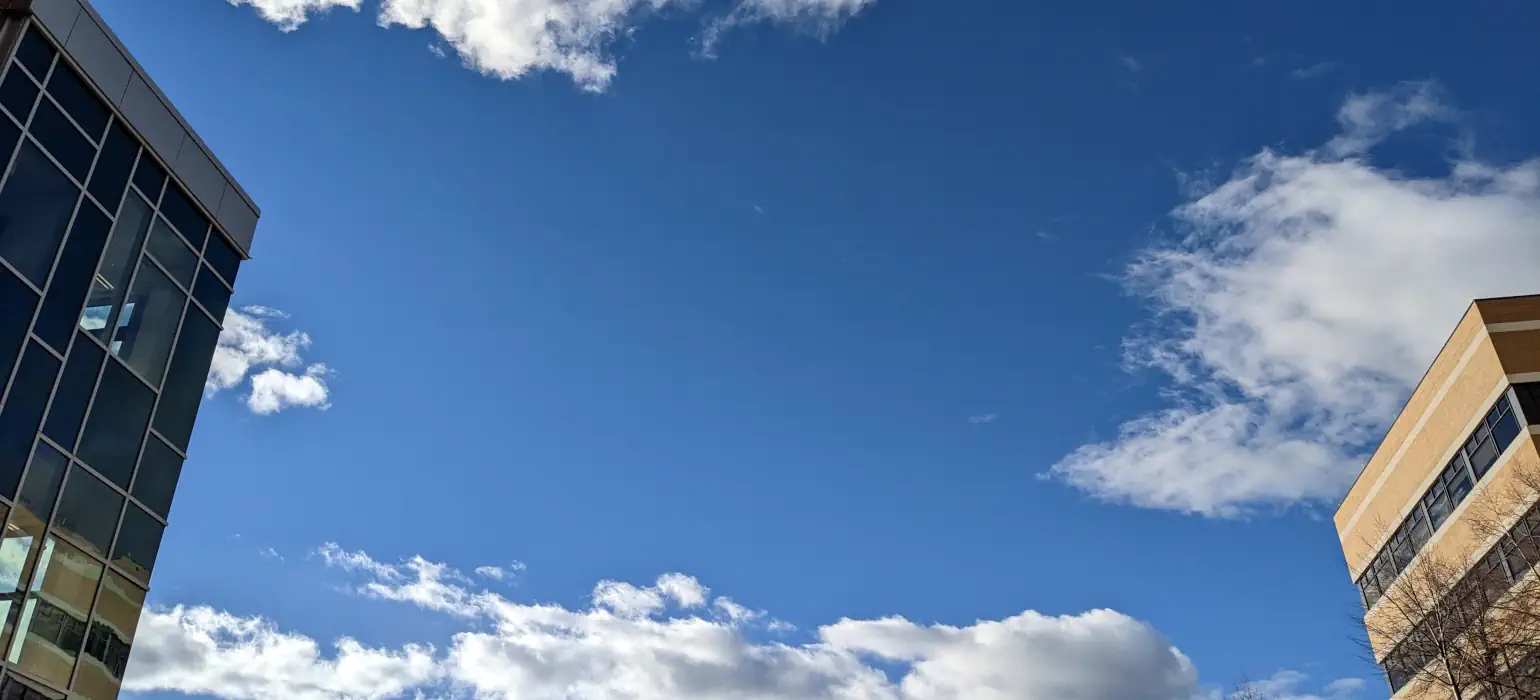 A blue sky with sparse clouds, framed by the tops of two buildings viewed from below.