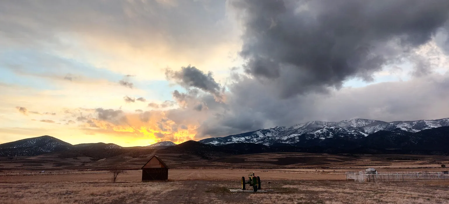 A fall sunset above an open plain: The yellow sun sets behind snowy mountains, casting orange glow on the left side of the sky, while the right side of the sky is dominated by dark storm clouds rising up and to the right in a dramatic diagonal. From the edge of the mountain towards the viewer spans a large brown plain. On it is a small shack and in the center of the image is a small, run-down tractor.