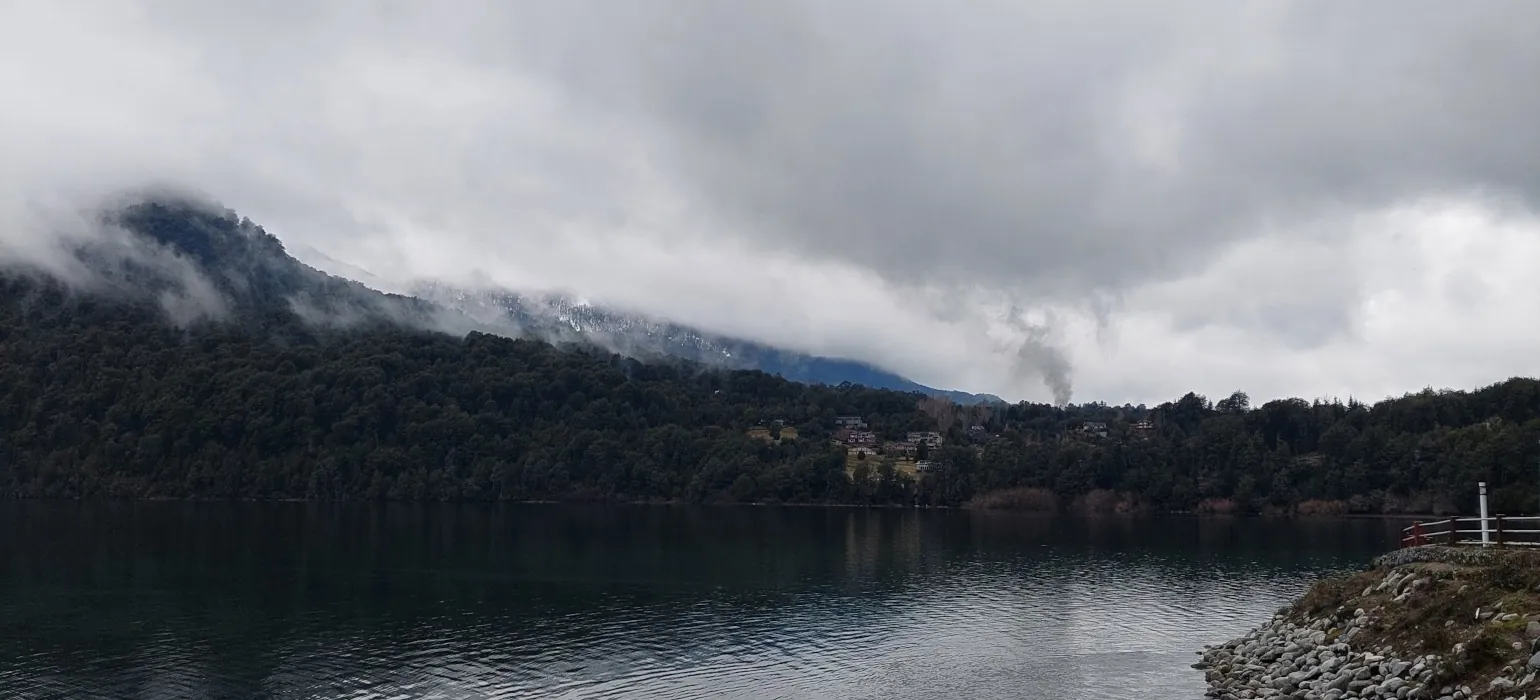 Looking across a lake, a mountain rises to the left, splitting the dark storm clouds from their reflection with a long, thin triangle of the mountain and its reflection. On the lower side of the hill is a small town, with its light green fields standing out against the otherwise unbroken dark green pine trees. The lake’s wind-blown ripply texture is broken up on the far right by a rock and grass outcropping, along with a railing on the right side.