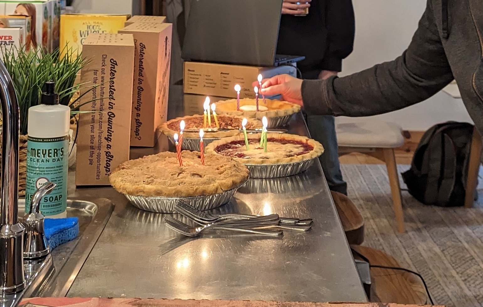 Four pies with lit candles in them sit on a steel countertop, as an End Pointer places the last candle.