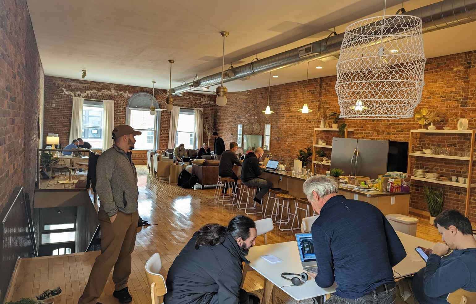 Casablanca. A wider view of the room, including the kitchen with a counter turned workstation. Several people are seen working in the foreground on a table, as well as the background on couches and other seats.