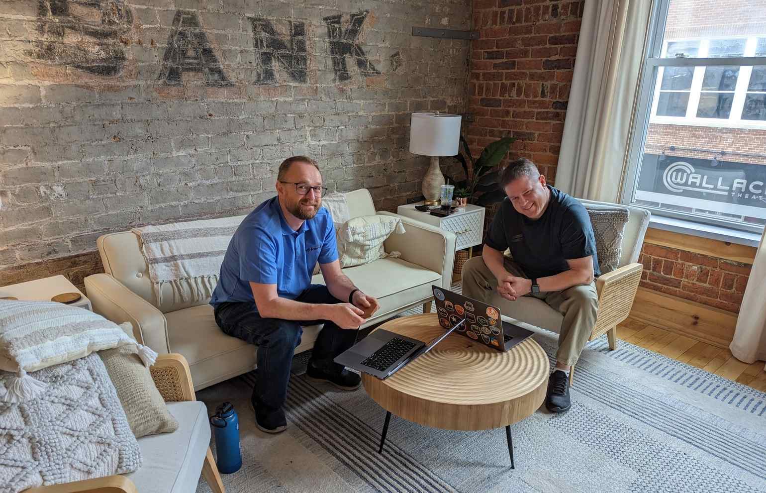 Casablanca. Another corner with a white couch and white seats, a modern wood table, and exposed brick on two walls. Two End Pointers are looking up from their laptops, smiling at the camera.