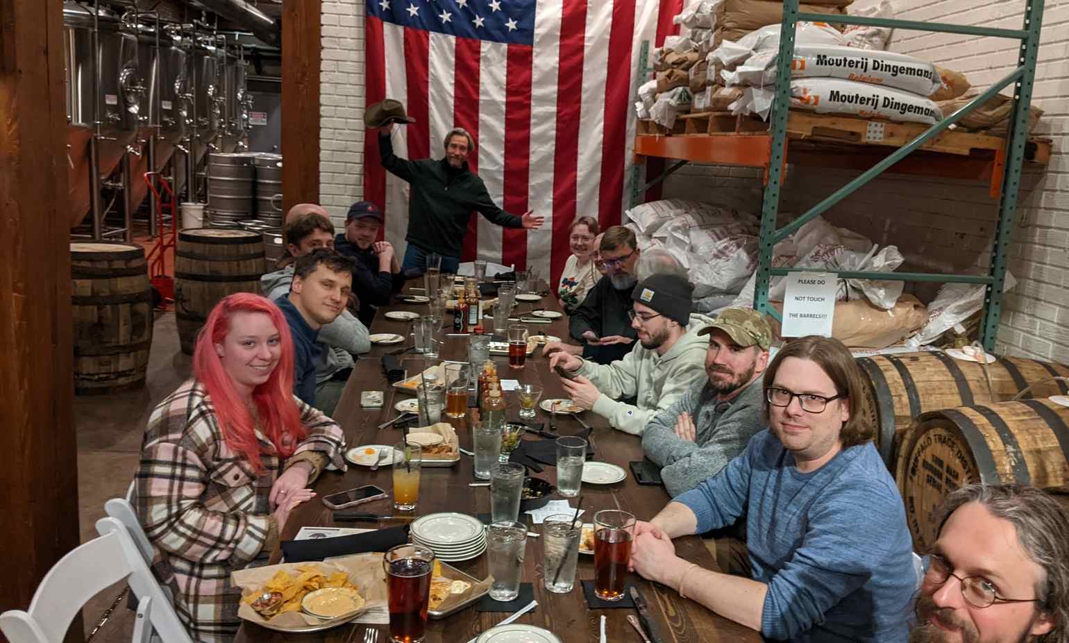 The opposite side of the same table. Ben waves with a cowboy hat from the back while other End Pointers smile at the camera, after the meal. Several barrels are visible around the restaurant/brewery.