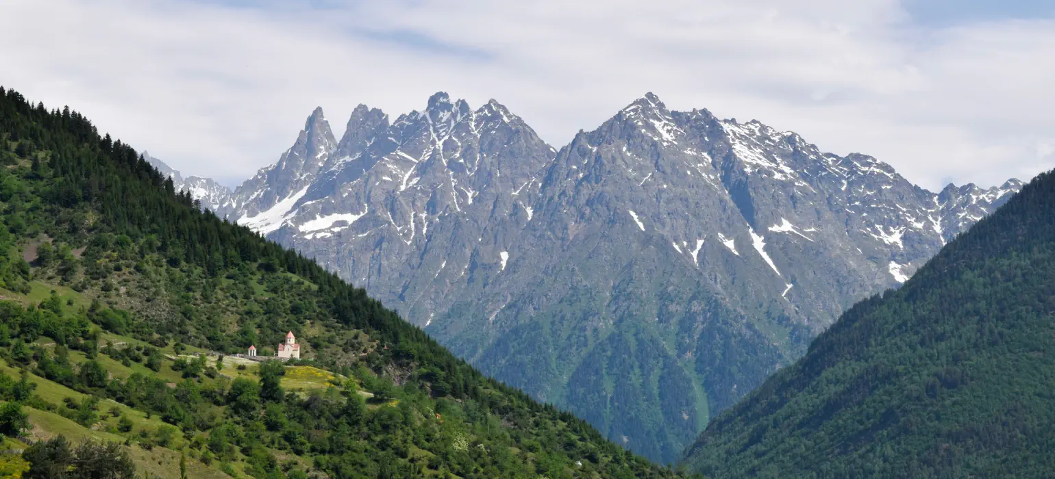 A steep, green mountain slope divides the image with a steep diagonal downwards and to the right, reaching the bottom of the frame two thirds of the way to the right edge of the image. A small, red-roofed Georgian church sits on it. Another hill sinks down and to the left, creating an apex at the bottom. In the background, framed by these asymmetrical diagonals, is a tall, rocky ridgeline lined with snow anywhere the rocks aren’t too steep to hold it.