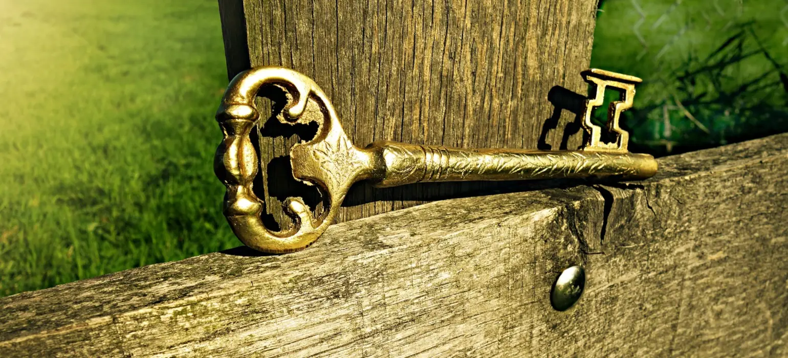 A close shot of a large golden key sitting on top of a wooden fence. There is green grass visible behind the fence, on the left and right upper sides of the image.