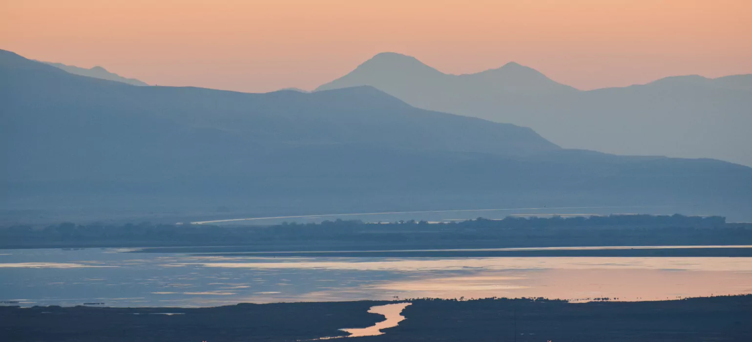 At sunset, the sky is a dark, fading orange above mountains which seen through layers of haze, making them appear blue. In the foreground, a lake with a large outcropping of land reflects the sky and mountains. At the bottom of the image much darker land is broken by a path of water travelling into the lake, reflecting orange, positioned directly into the center of the frame.