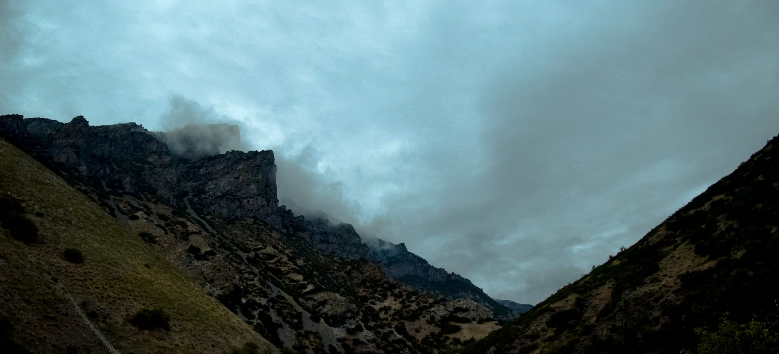 A dark sky sprawls over a tall canyon. Misty clouds hang on jagged peaks on the hill to the left. The other hill on the right rises at a steep angle, making a “V” shape. Both hills are covered in different shades of green, mostly a deep, dark green due to the late hour. Some sun peeks through the overcast sky to let through a bit of bluer light.