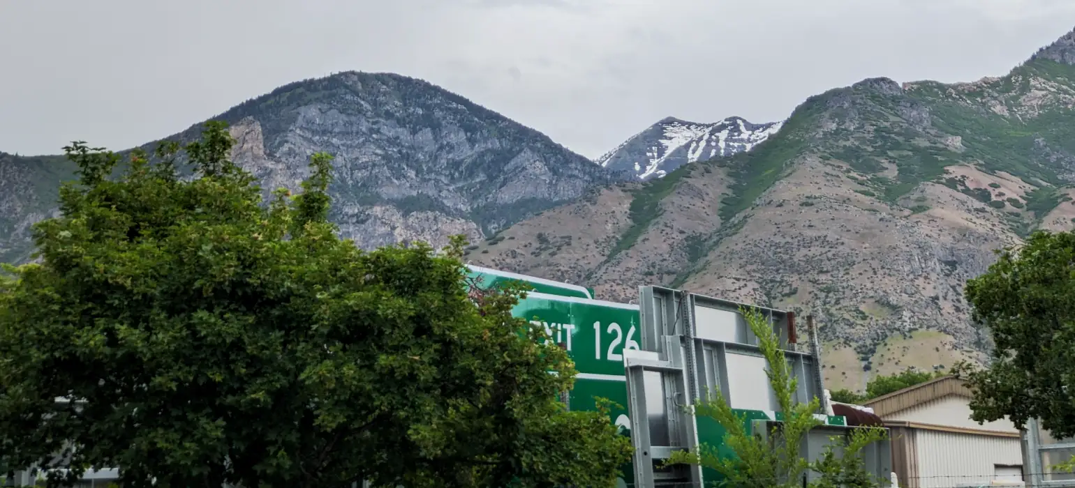 A nearby mountain skyline lies against an overcast sky. The lower hills are obscured on the edges by close green trees in the foreground, which lead toward US freeway signs in storage.