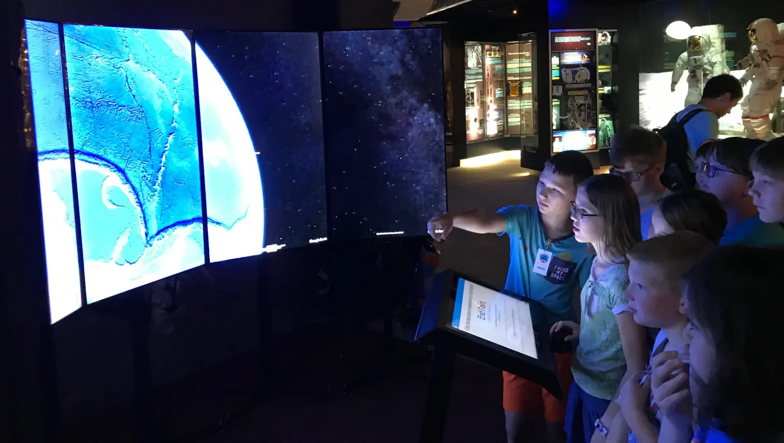 Several school kids gather around a VisionPort installation in a space museum, with spacesuits visible in the background. A boy points at an image of a planet displayed on the seven screens arranged in a semicircle.
