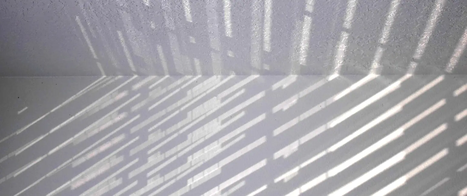 A blank white wall and ceiling, with regular rays of light casting striped shadows at an acute angle. As the light hits the ceiling, the angle changes and the strips of light appear vertical.