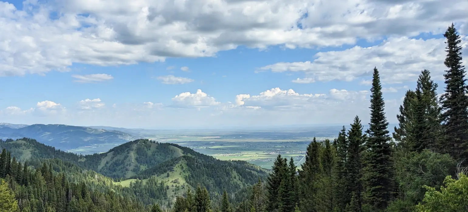 The view from a mountain ridge. the sky is light blue and partially covered in clouds. Green ridges covered in pine trees lead down to a flat valley populated by a small town.