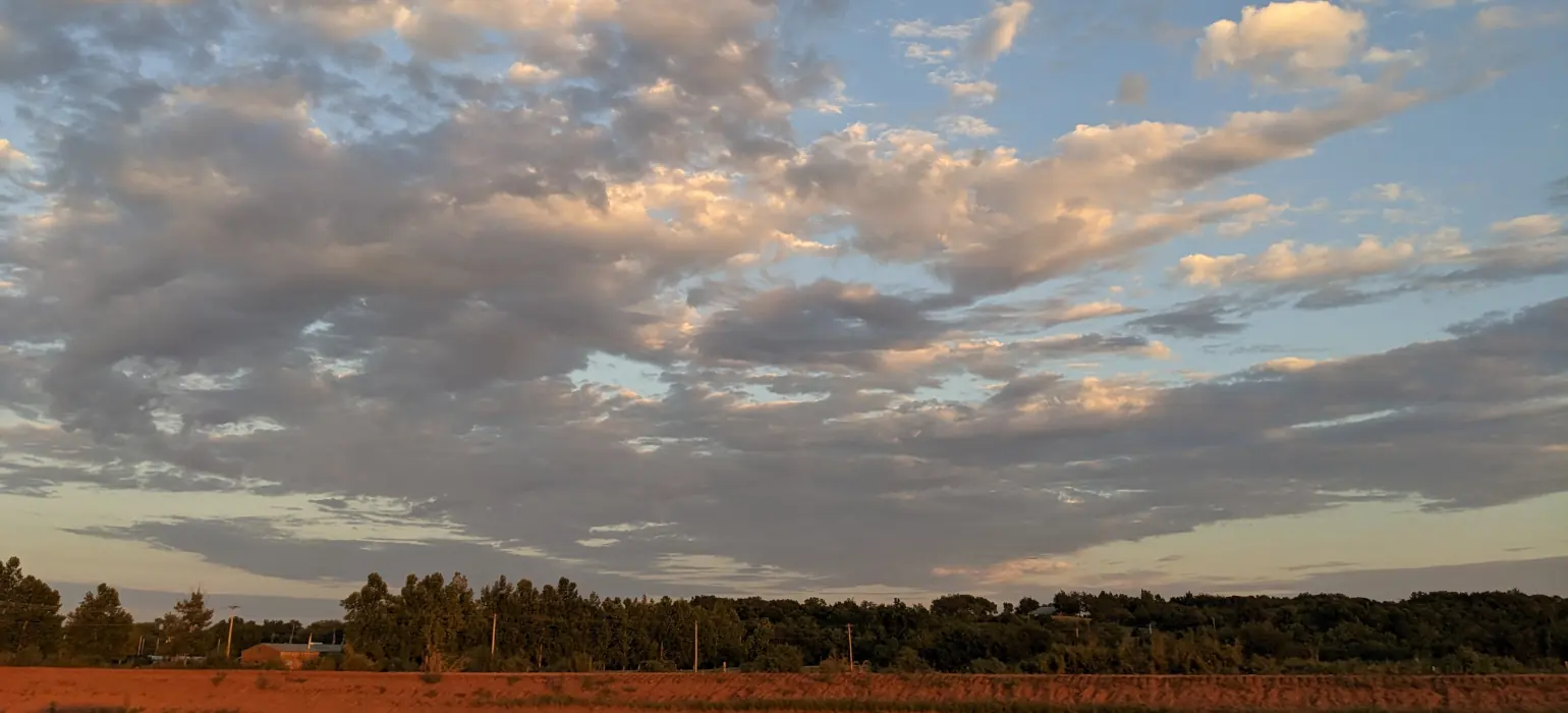 An expansive sky filled with faintly red clouds extends above a field turned red from the sunset. A layer of trees separates the field from the sky