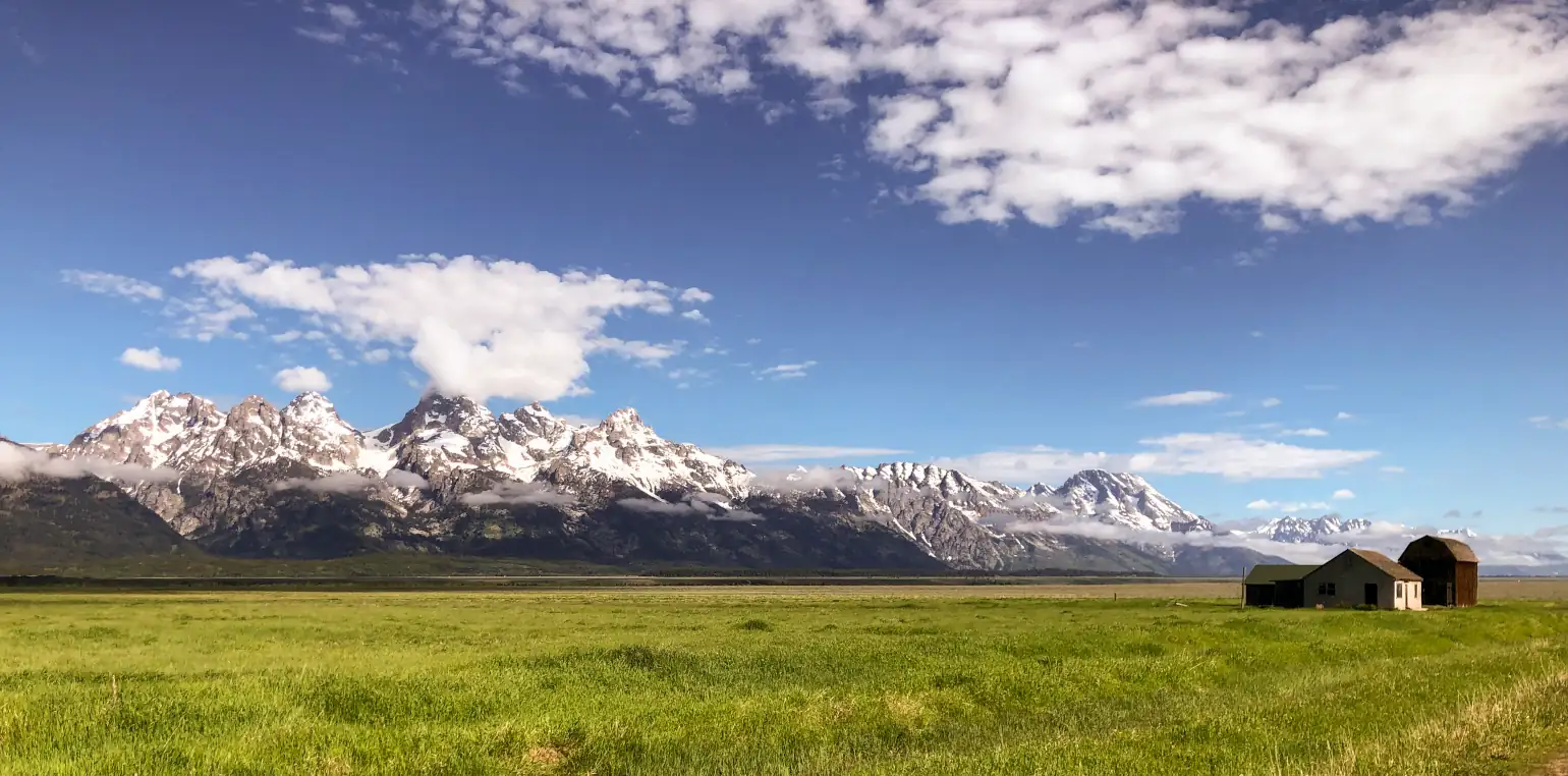 Photo taken of and from the Mormon Row Homestead at Grand Teton National Park. Photo is taken facing WNW, and has an old home and barn in the foreground with the jagged, rocky peaks of the Teton mountain range in the background.