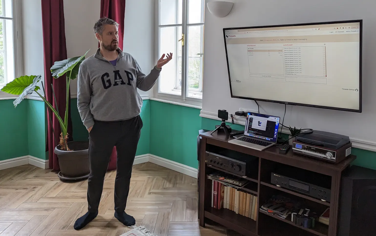 Man in an apartment looks toward large TV screen mounted on a wall, showing a computer display of a website