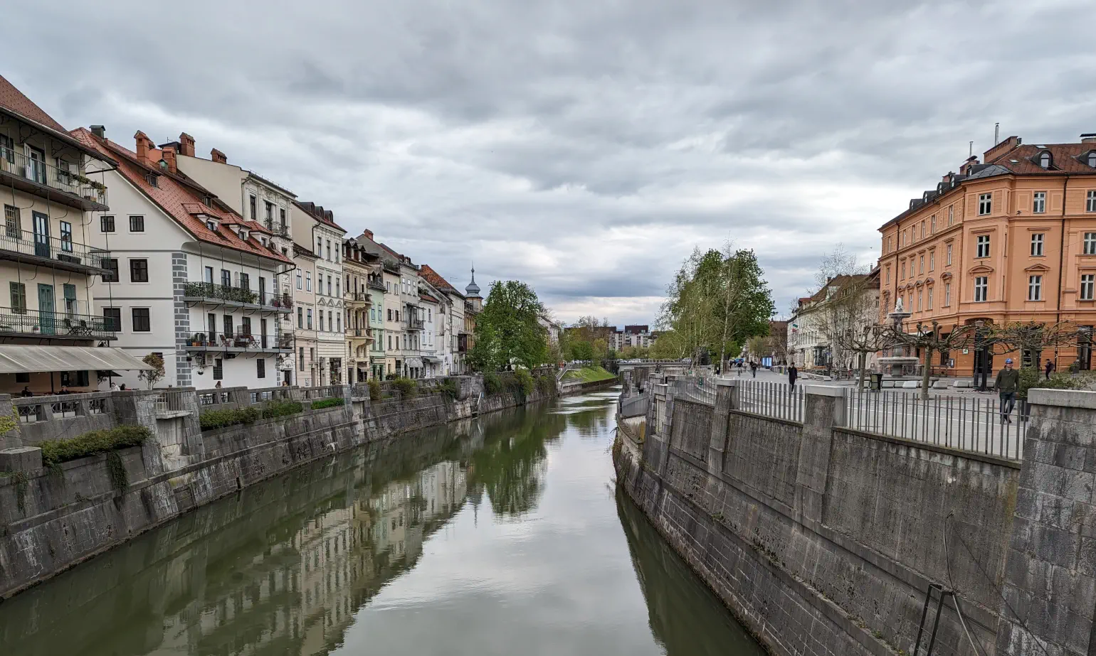 Colorful 4-story buildings with trees along concrete city walls containing a river reflecting a cloudscape