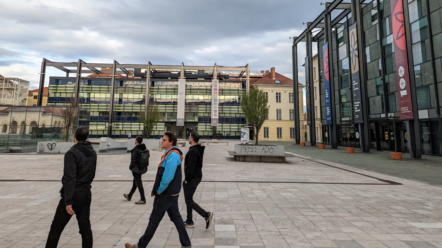 4 men walking left across a city square in front of buildings with modern architecture