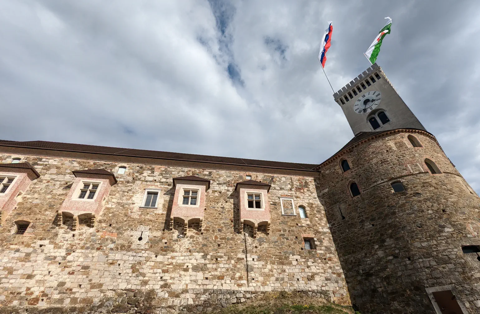 A sunny view of a castle wall and tower with flags flying in the breeze, against a blue sky with white clouds