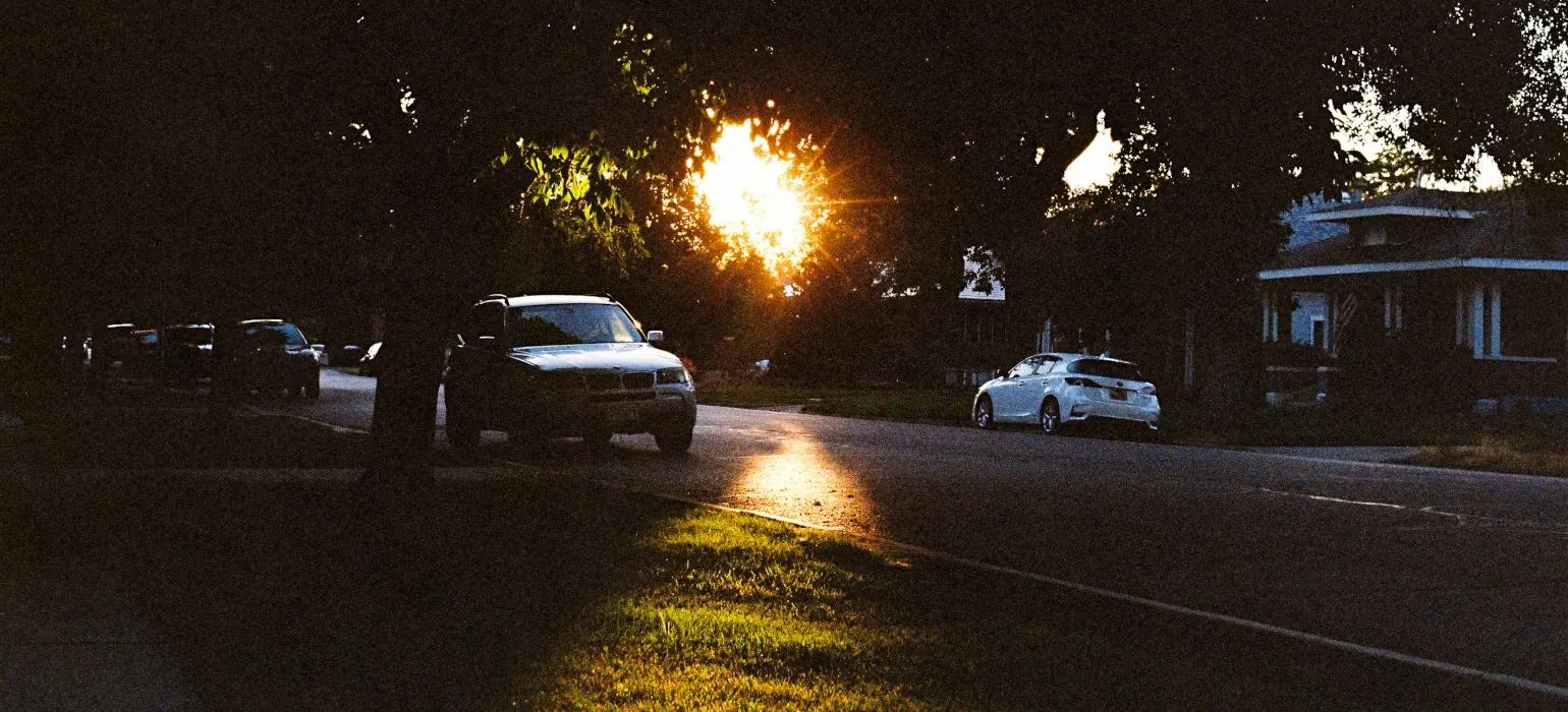 In the center of a dim street lined with houses and cars, the sun illuminates and sillhouettes green foliage. It projects a triangle of light on the ground, first the street and then the grass, centered in the frame. On either side of the light, centered verticaly, a white car catches the light.