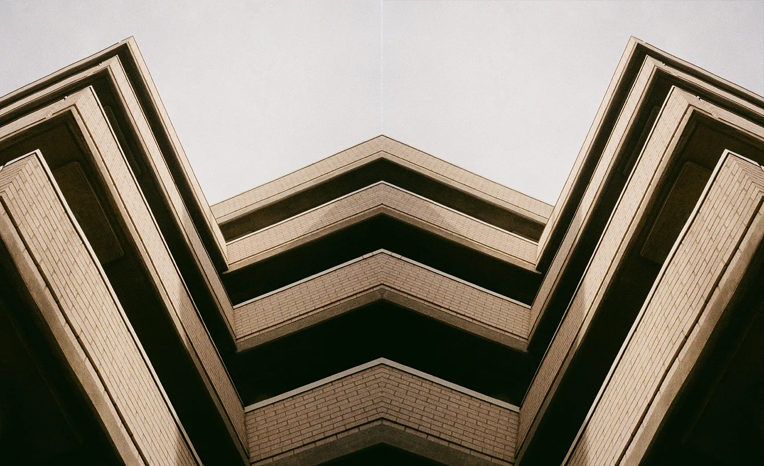 A low-angle view of a brick building with half height walls creating an alternating pattern of wall and dark, shaded ceiling. mirrored about the center of the image. Each mirrored side of the image has two strong angles; the building protrudes from midway up the left edge, turning sharply down, then coming back up and to the right to the center.