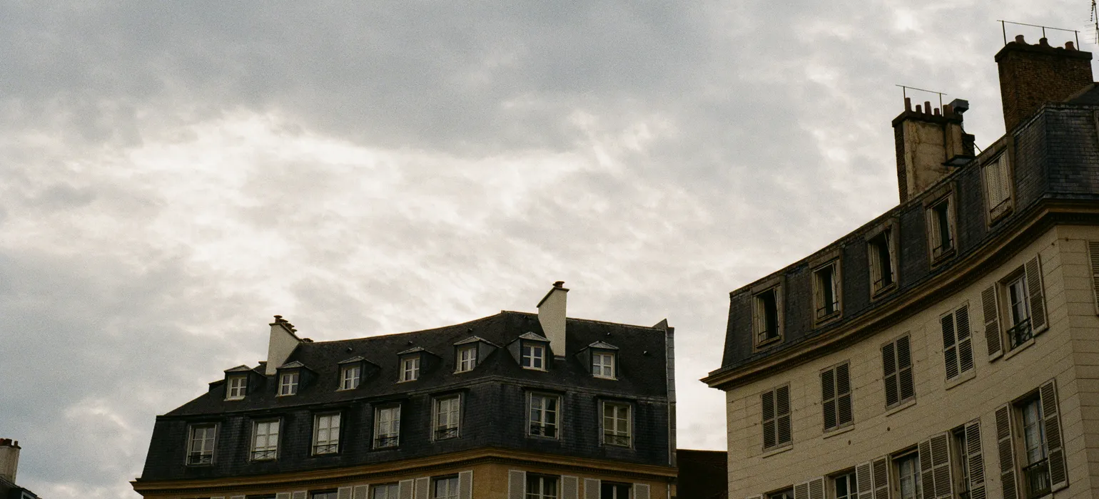 European rooftops curve at the bottom of the image upwards. The rest of the image is a partially overcast gray sky with some sun peeking from behind the clouds