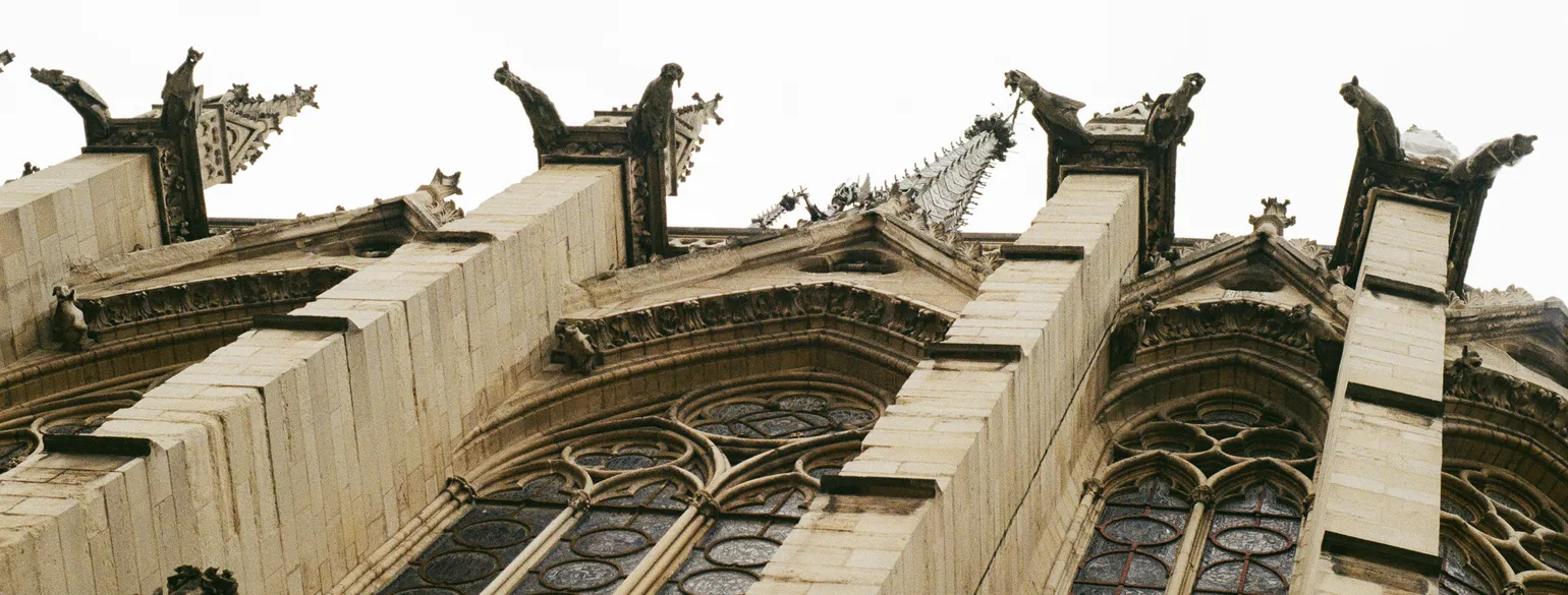 The side of a gothic church from a very low angle. The ornate spires and gargoyles loom over the viewer, with dark stained glass beneath, sectioned by square stone pillars.