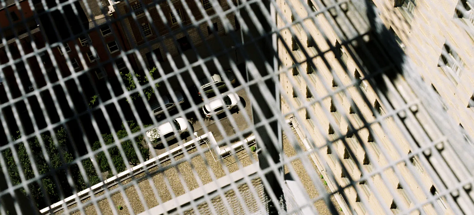 Through the metal grid of a fire escape step, cars and a street far below are visible. On the step are two strong intersecting shadows from the top left, one going rightward and one going downward and to the left
