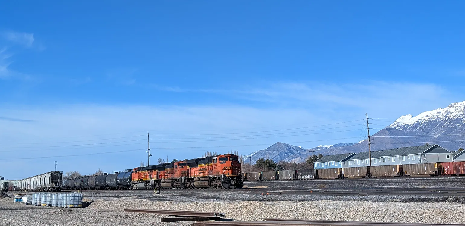 A long freight train points to the right, slightly toward the camera. The train cars extend off the image to the left. Above the tracks is a white snow-capped mountain and a deep blue sky.
