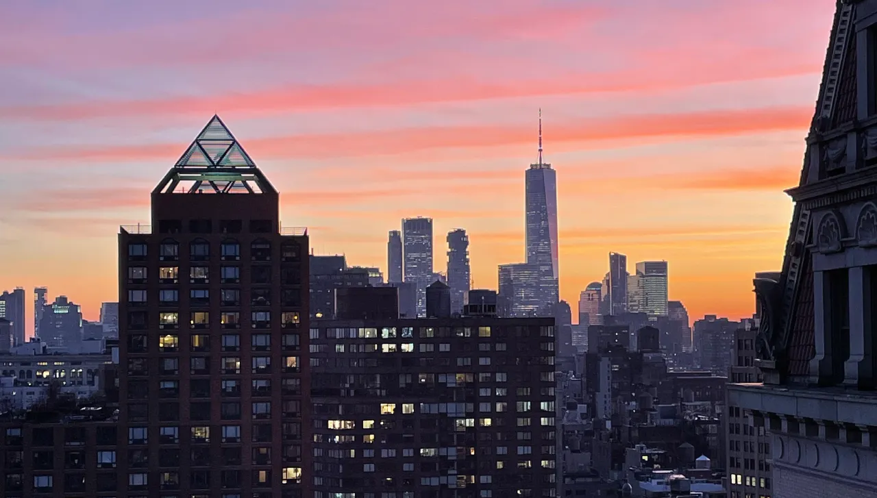 New York City skyline at dusk, with warm orange and pink hues across the sky and a foreground building topped with a glass pyramid structure.