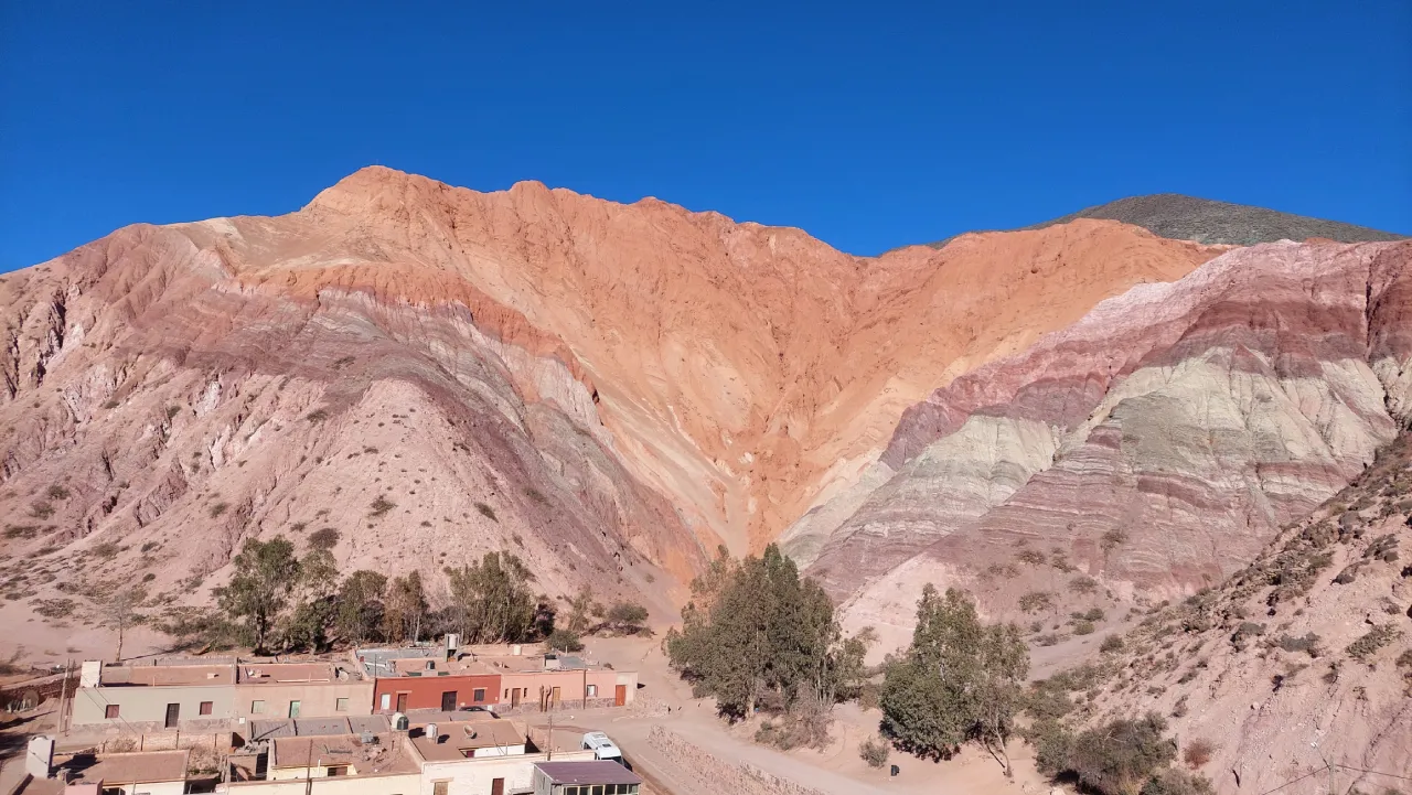 A striped, multicolored mountainside rising above a small adobe village under a deep blue sky