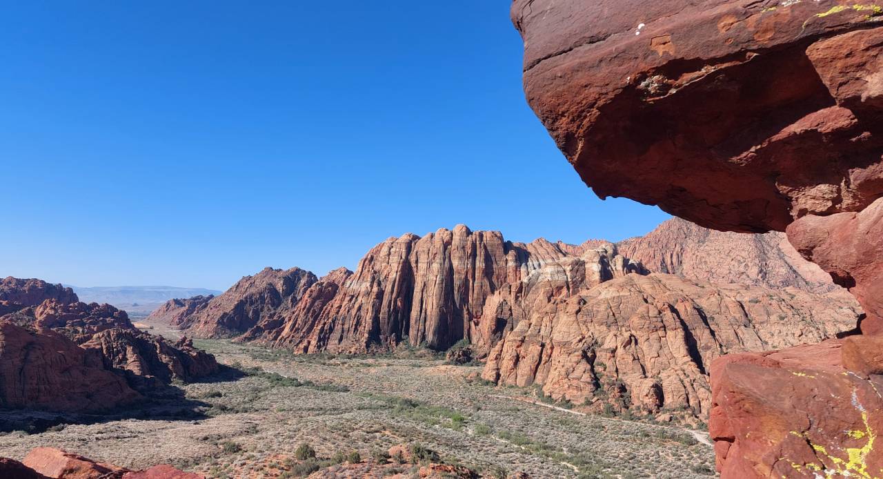 A desert valley lined with tall red and cream sandstone cliffs under a clear blue sky, with a red rock overhang in the foreground.